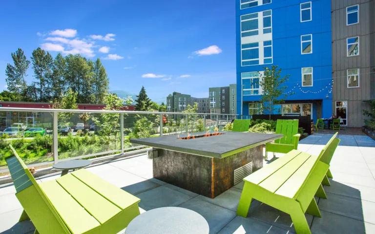 Rooftop patio area with a fire pit, surrounded by green wooden benches and a view of nearby buildings and trees.