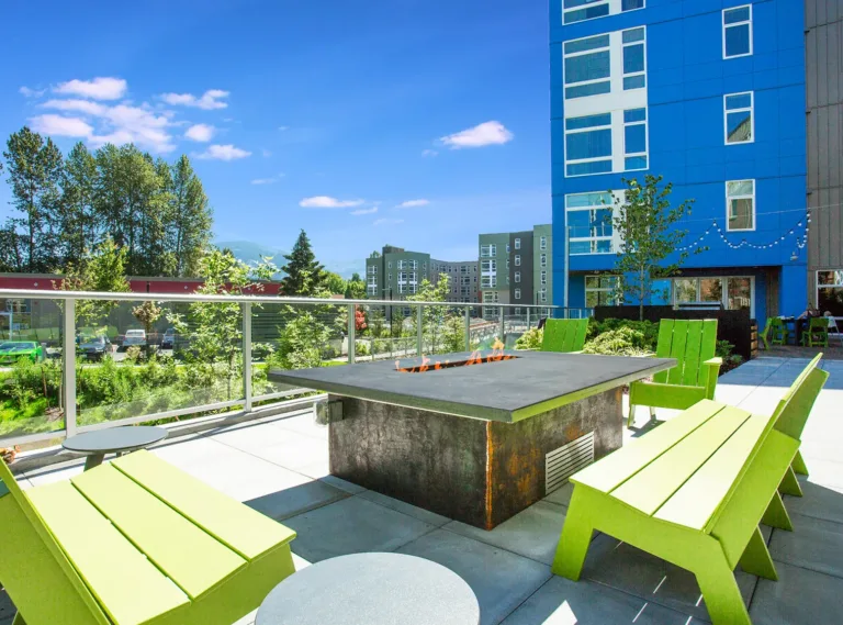 Rooftop patio area with a fire pit, surrounded by green wooden benches and a view of nearby buildings and trees.