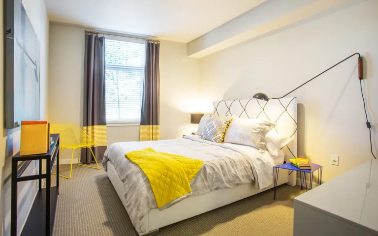 Apartment bedroom with a modern design, featuring a double bed, colorful accents, and large window with natural light.