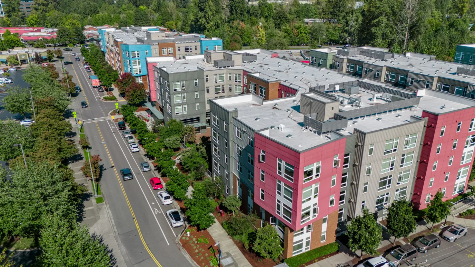 Aerial view of modern apartment buildings featuring colorful facades in shades of red, blue, and beige, with a tree-lined street running alongside and cars parked on both sides.