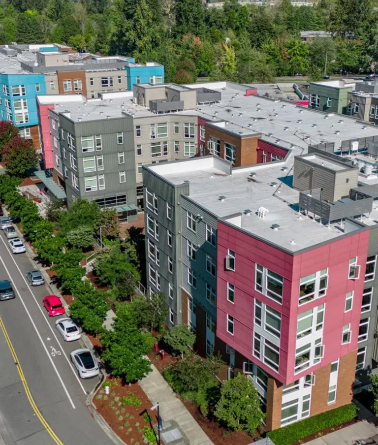 Aerial view of modern apartment buildings featuring colorful facades in shades of red, blue, and beige, with a tree-lined street running alongside and cars parked on both sides.
