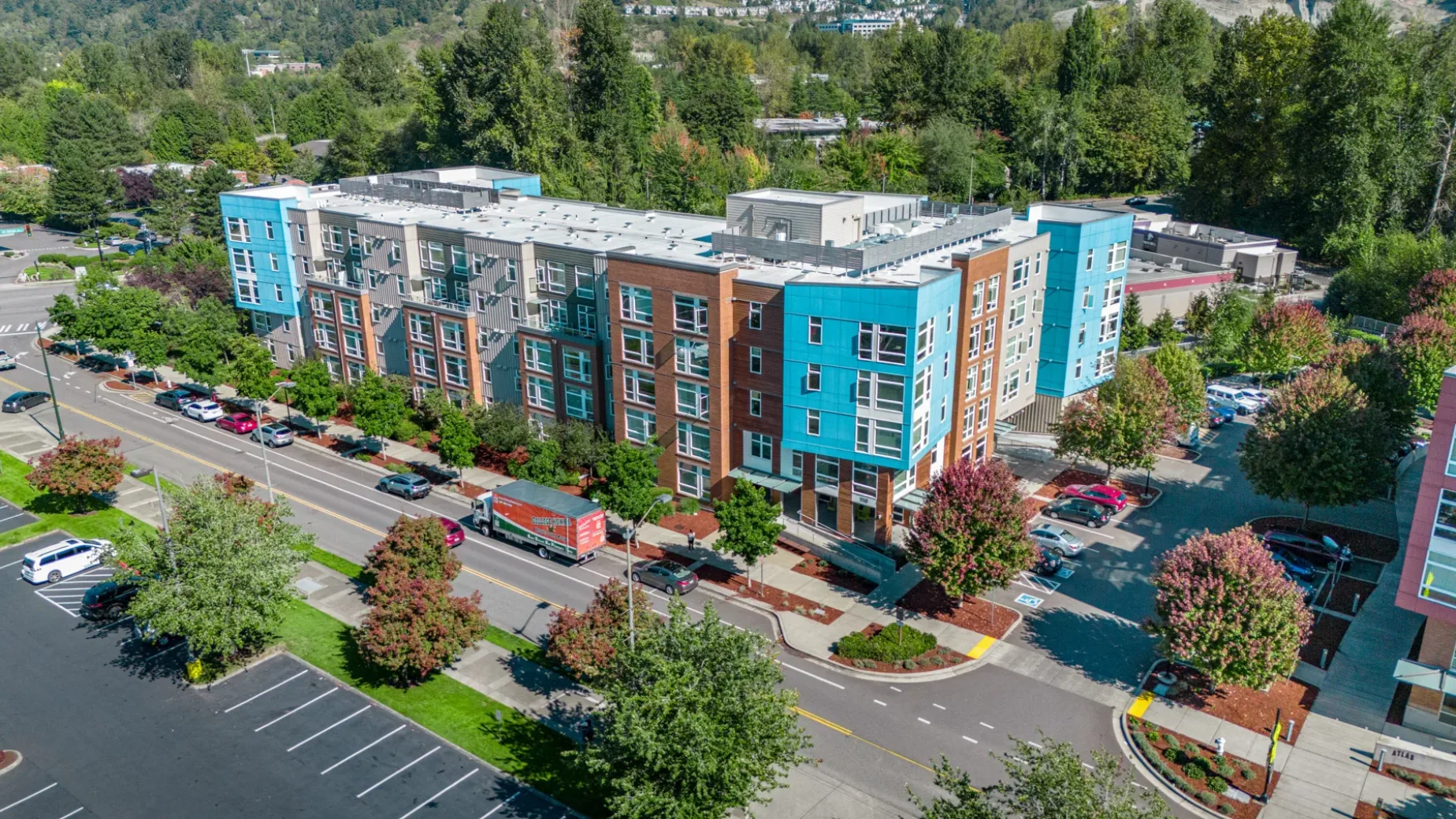 An aerial view of a modern apartment complex with colorful facades featuring blue and beige panels, surrounded by trees and parking spaces. The road near the complex has parked cars and a delivery truck.