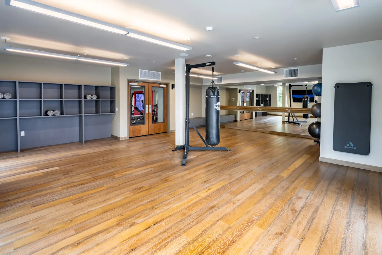 A spacious gym area with wooden floors, a punching bag, shelves with towels, and exercise balls in the corner. Large mirrors cover one wall and bright lighting is overhead.