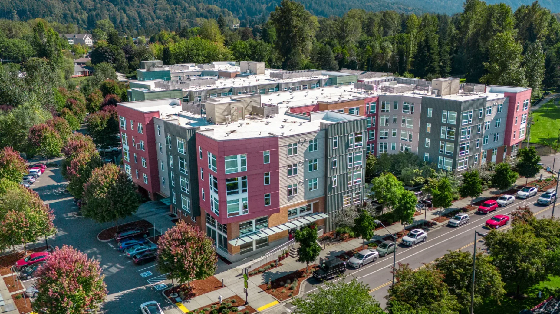Aerial view of a modern apartment complex with colorful facades, surrounded by trees and lined with parked cars on the streets.