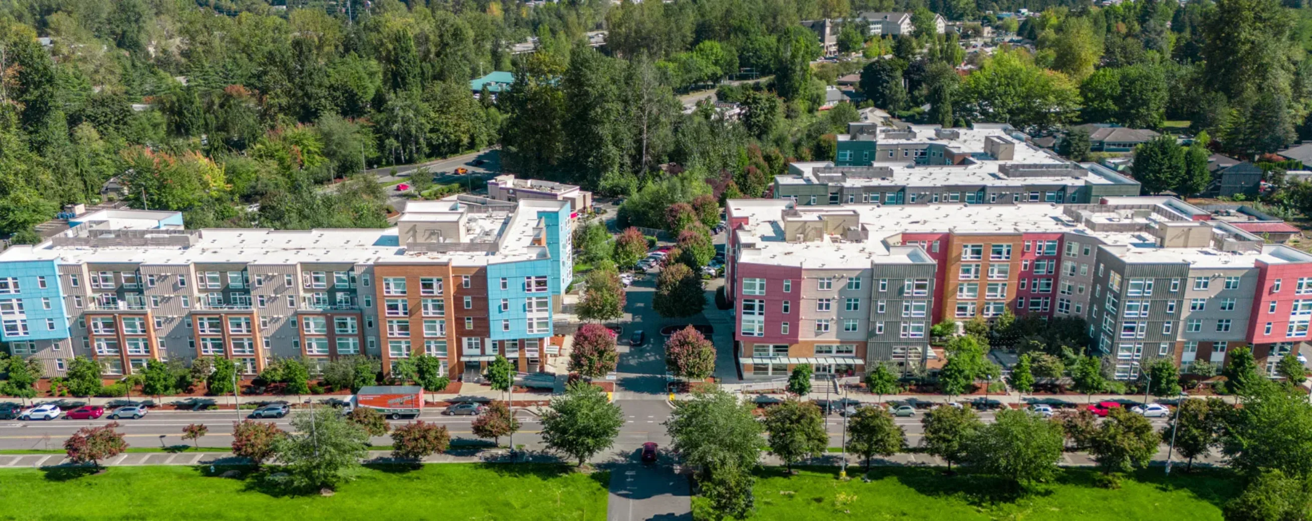 Aerial view of multi-story apartment buildings surrounded by trees and greenery, with a road and parked cars in front.