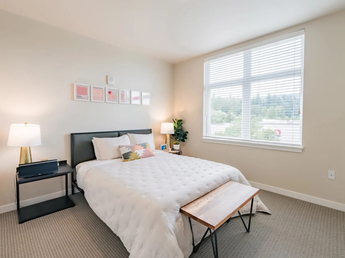 Modern apartment bedroom with a neatly made bed, large window with blinds, and decorative lamps on each side.