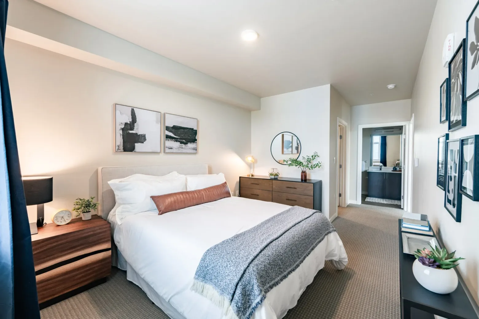 A modern apartment bedroom featuring a neatly made bed with white linens, wooden furniture, abstract wall art, and a small plant, leading to a bathroom.