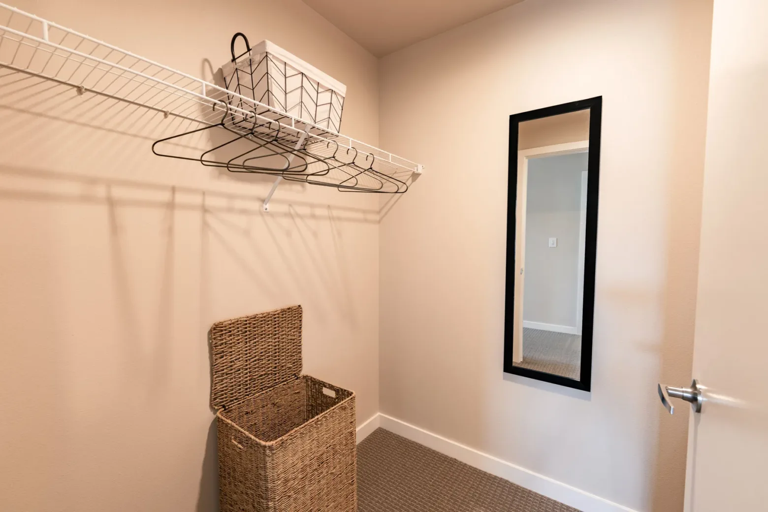 Apartment closet with a wire shelf holding hangers and a basket, alongside a full-length mirror and a woven hamper on the floor.