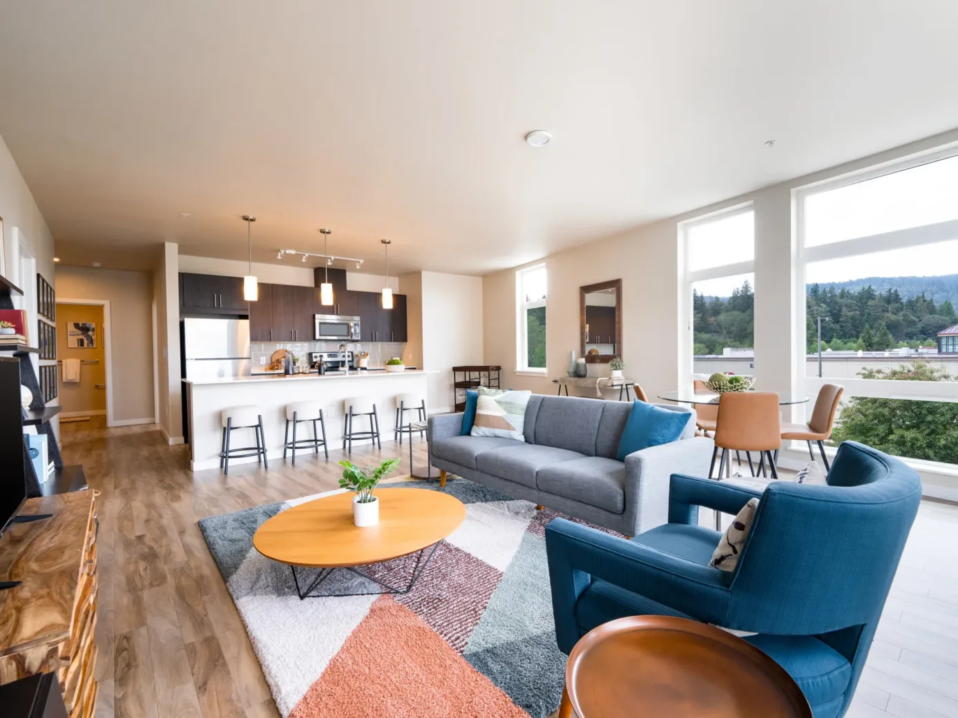 Spacious apartment living room with wooden floors, a blue armchair, a gray sofa, and a dining area with large windows overlooking a scenic view.