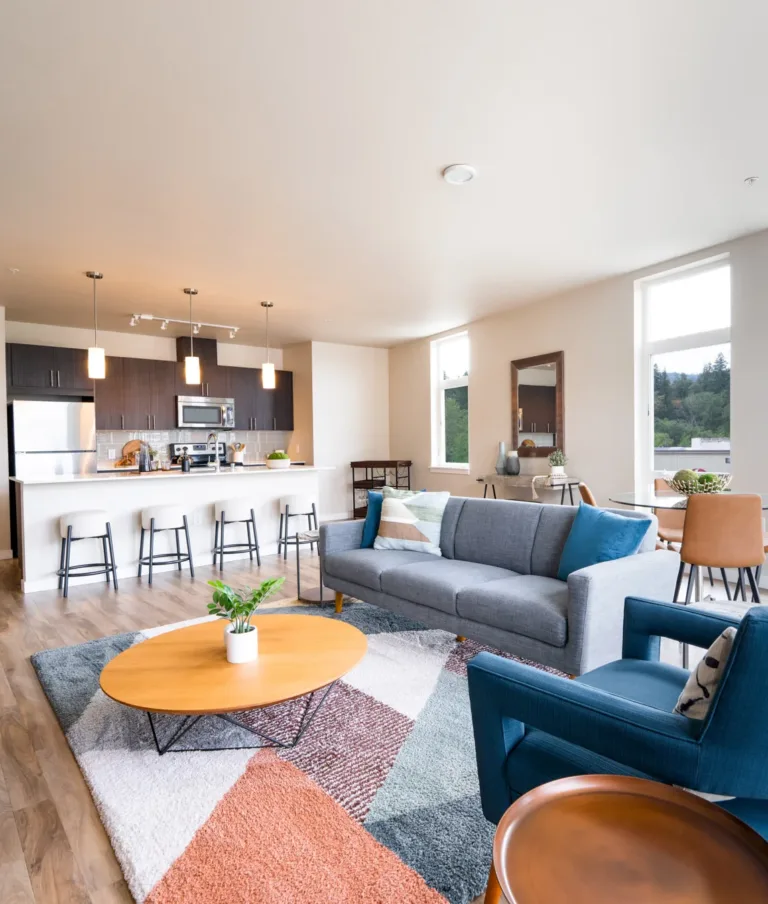 Spacious apartment living room with wooden floors, a blue armchair, a gray sofa, and a dining area with large windows overlooking a scenic view.