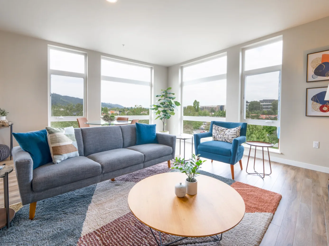 Modern apartment living room with a gray sofa, blue armchair, colorful rug, and large windows offering a scenic view.