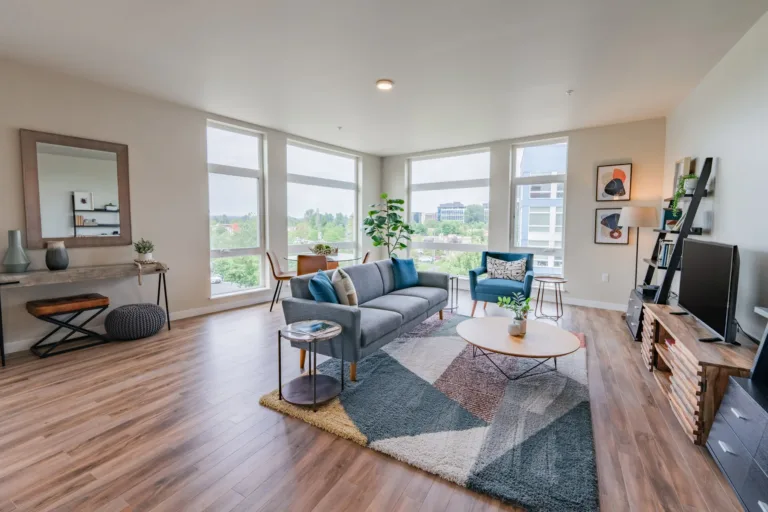 Modern apartment living room with a gray sofa, colorful decor, and large windows offering a city view.