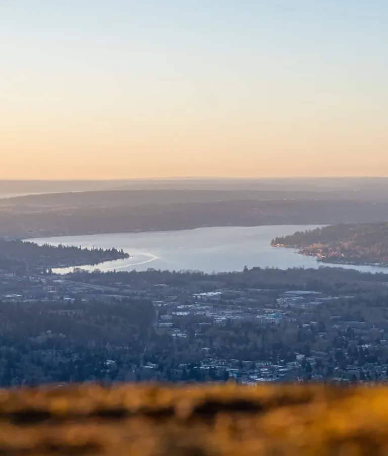 A scenic view of a Lake Sammamish surrounded by forested hills and scattered urban areas, under a gradient sky transitioning from orange to blue, indicating sunset or sunrise.
