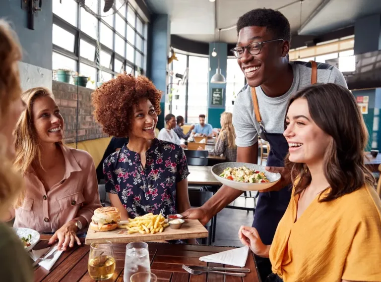 A server in glasses and an apron serves a smiling group of three women at a restaurant, each with their own meal in front of them. The atmosphere is lively, with other diners visible in the background.