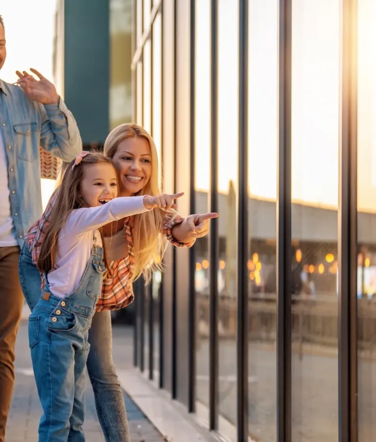 A family with a man, woman, and young girl pointing and smiling while looking through a glass window, holding shopping bags under a warm, glowing sunset.